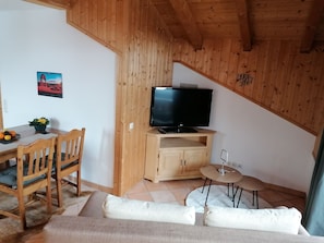 A cozy room with a wooden ceiling, a TV on a wooden stand, a dining table with chairs, a sofa, and a small round table with a lamp.