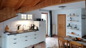 Una cocina con armarios blancos, encimera negra y techo de madera. Hay una zona de comedor con una mesa y sillas, y una estantería con objetos decorativos.