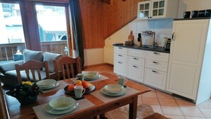 Un comedor con una mesa de madera dispuesta para cuatro personas, rodeada de sillas. Se ve una cocina con armarios blancos y una encimera al fondo.