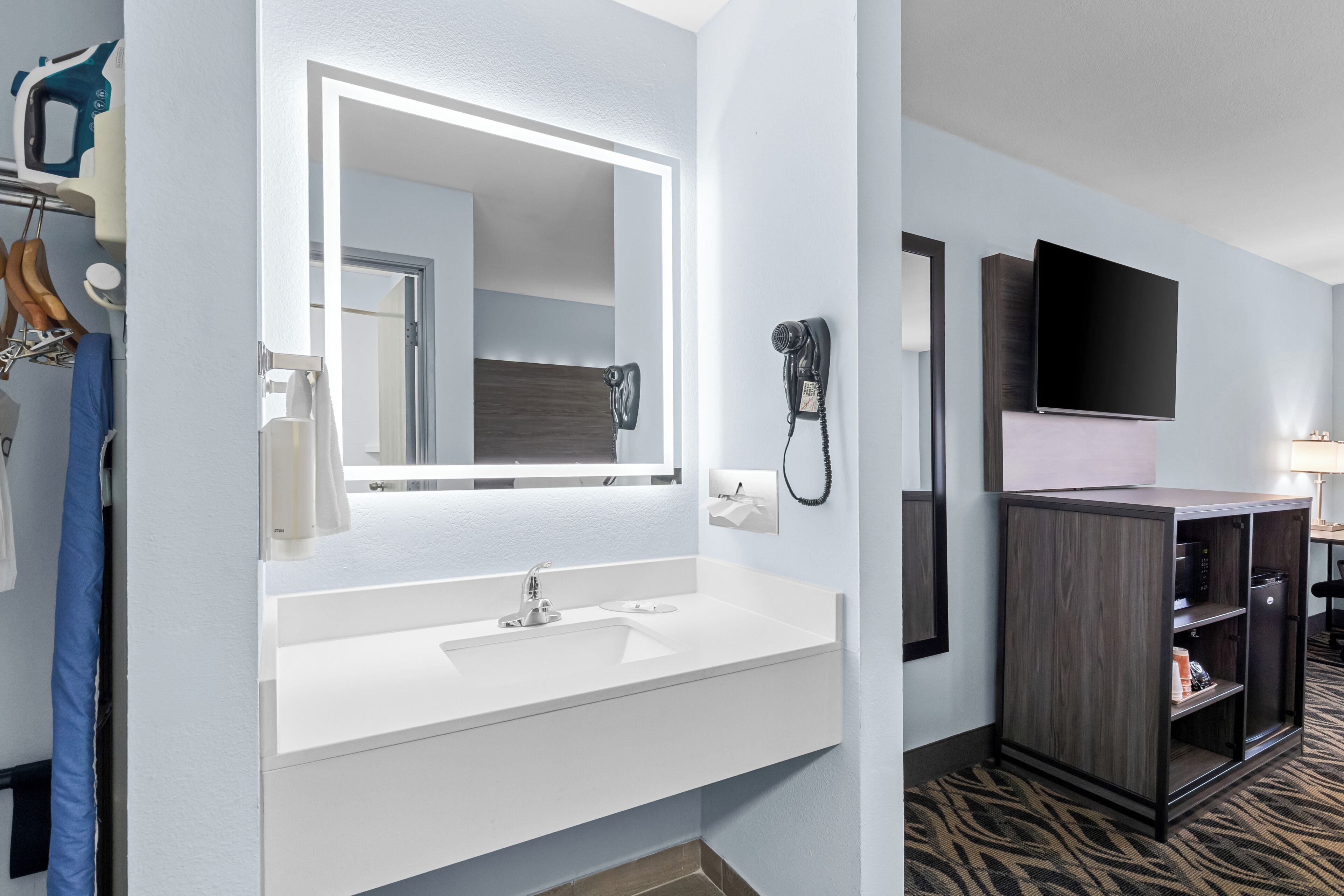 A hotel bathroom with a white sink, a large mirror, a hairdryer, and a television mounted on the wall.