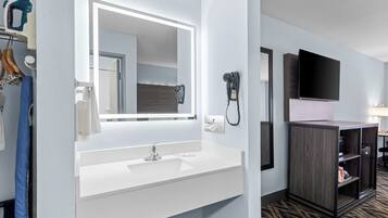 A hotel bathroom with a white sink, a large mirror, a hairdryer, and a television mounted on the wall.