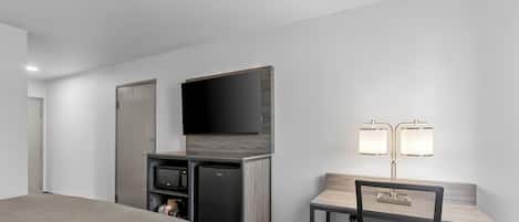 A hotel room with a bed, a desk with a chair, a television, a minibar, and a lamp.