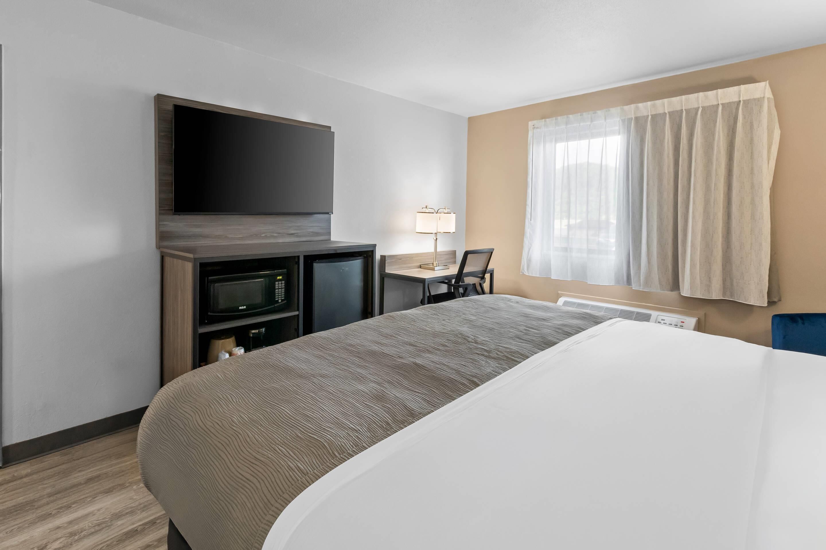 Ein Hotelzimmer mit einem Bett, einem Flachbildfernseher, einer Mikrowelle, einem kleinen Schreibtisch mit Lampe und einem Fenster mit Vorhängen.