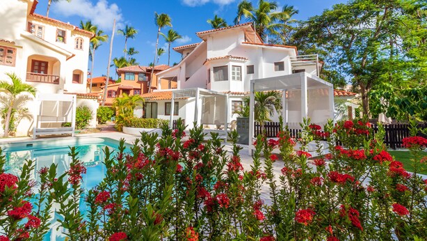 A resort with a swimming pool, palm trees, and a white building with a red roof. - Amazing Villa Bavaro Beach (Punta Cana)