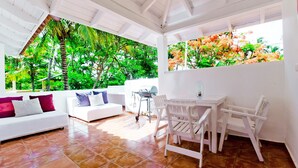 A bright living room with a white sofa, a dining table with chairs, and a view of lush greenery through large windows. - Amazing Villa Bavaro Beach (Punta Cana)