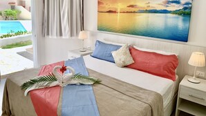 A hotel room with a bed, a nightstand, a lamp, a painting of a sunset over a lake, and a view of a swimming pool outside. - Amazing Villa Bavaro Beach (Punta Cana)