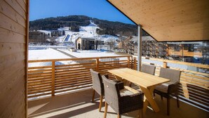 Un terrazzo in legno con tavolo e sedie, affacciato su un paesaggio innevato con edifici e una montagna all'orizzonte.