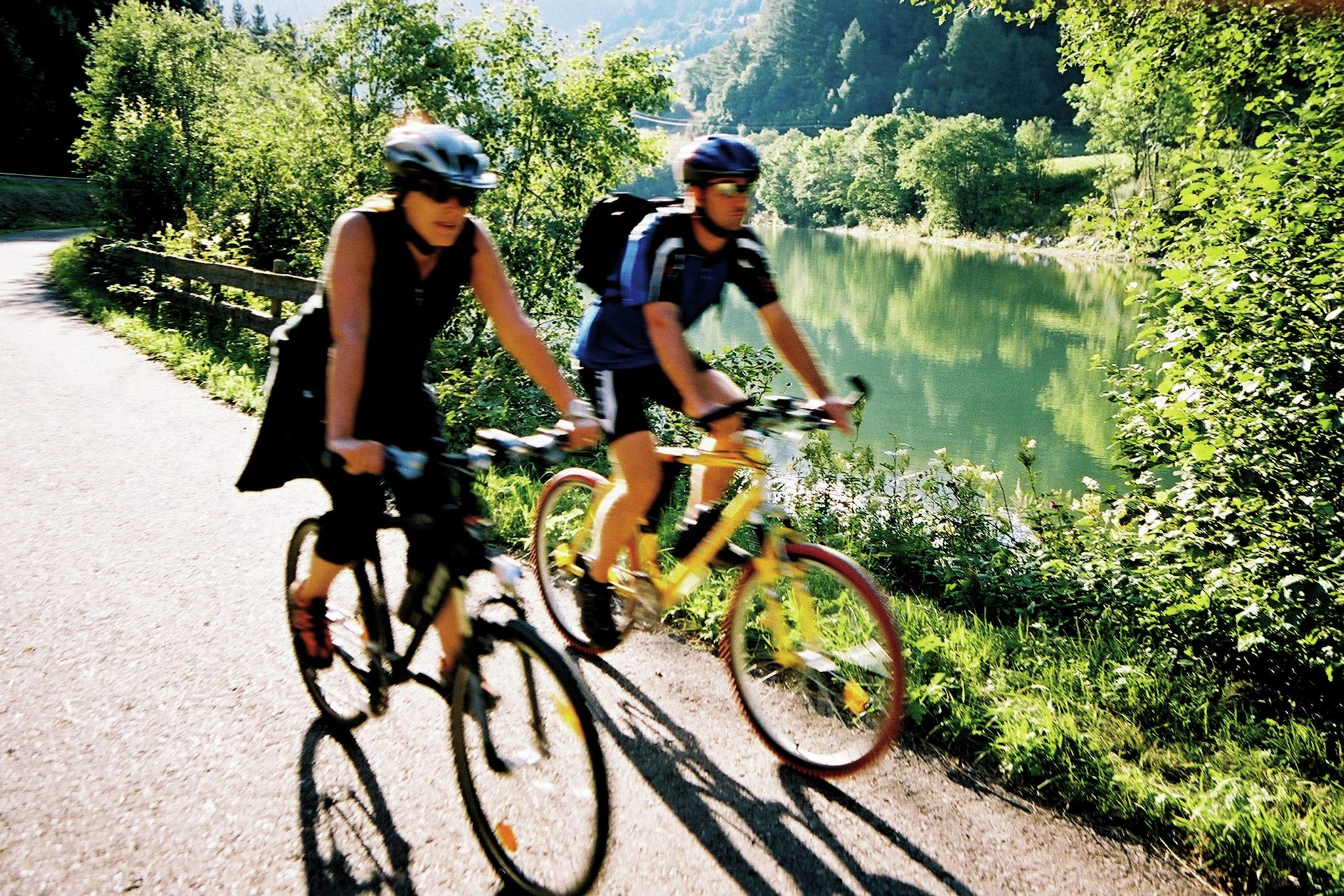 Two cyclists on a paved path beside a lake, surrounded by greenery.
