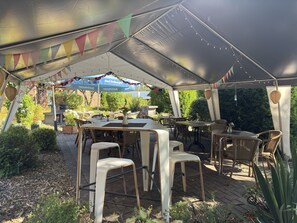 Une terrasse extérieure avec des tables et des chaises, entourée de verdure et dotée d’un espace couvert avec une enseigne.
