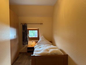 A small, narrow room with a single bed, a nightstand, a window with curtains, and a wooden wardrobe.