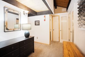 A room with a wooden ceiling, a black dresser, a mirror, a lamp, a framed picture, and a door. - Sundowner Mountain View Condo • Walk to Slopes & Main St  • Pool Access (Breckenridge)