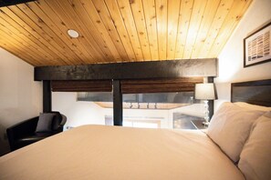 A hotel room with a wooden ceiling, a bed with white bedding, a desk with a lamp, and a view of the outside. - Sundowner Mountain View Condo • Walk to Slopes & Main St  • Pool Access (Breckenridge)