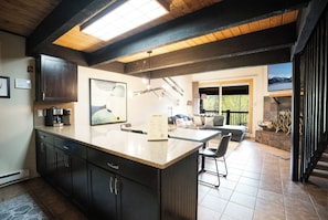 A modern kitchen with dark cabinets, a large island, and a dining area with a view of the outdoors. - Sundowner Mountain View Condo • Walk to Slopes & Main St • Pool Access (Breckenridge)