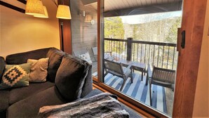 En hyggelig stue med en mørk sofa, dekorative puder og en stor skydedør, der fører ud til en terrasse med udsigt over træer og en klar himmel.