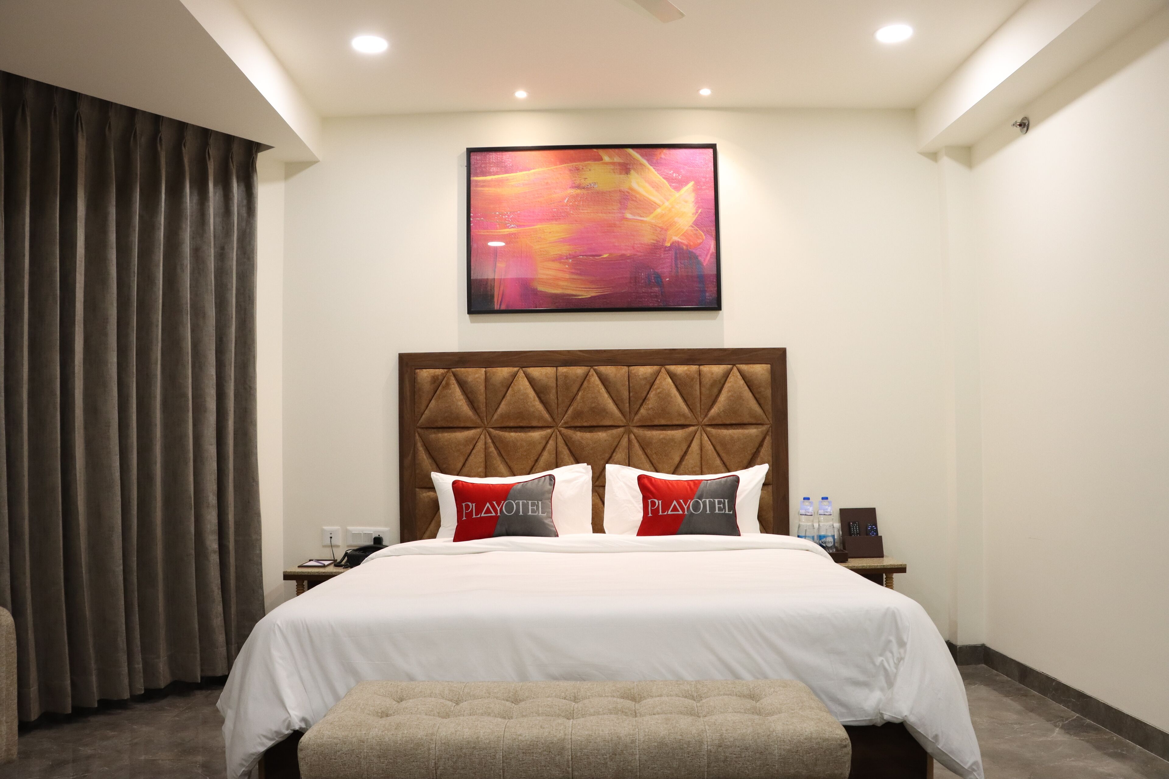 Une chambre d’hôtel avec un grand lit, une tête de lit ornée d’un motif géométrique, deux oreillers portant le logo Playotel, une table de chevet avec un téléphone et des bouteilles d’eau, et un grand tableau abstrait accroché au mur.