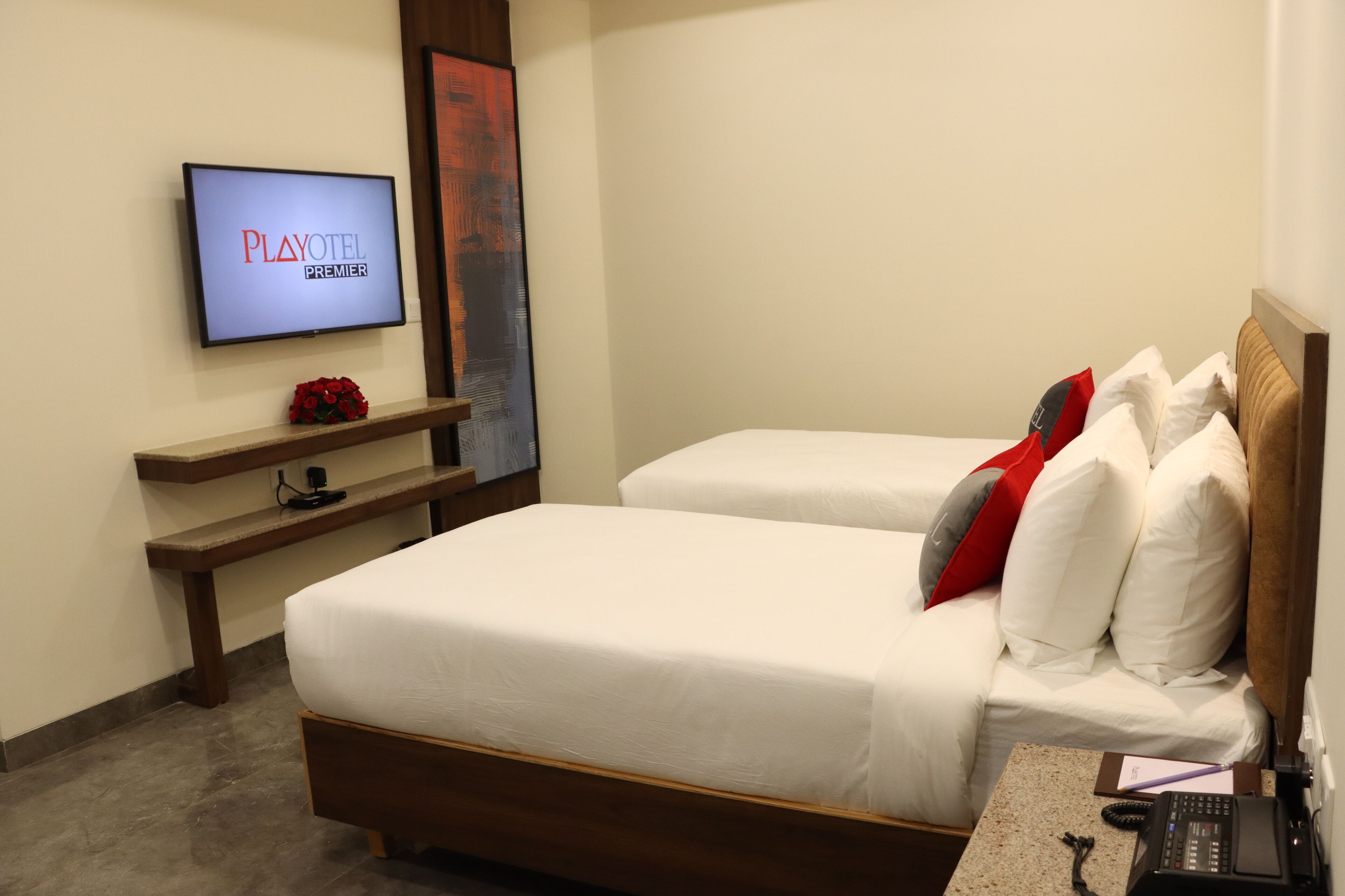 A hotel room with a bed, a TV mounted on the wall, a small desk with a phone, and a red decorative item on the shelf.