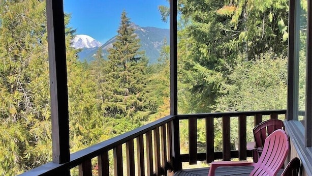 A wooden deck with two Adirondack chairs, overlooking a forest and a mountain.
