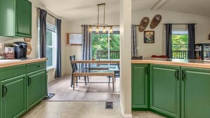 Een keuken met groene kastjes, een eettafel en een bankje. Er is een raam met gordijnen, een hanglamp en uitzicht op groen buiten.