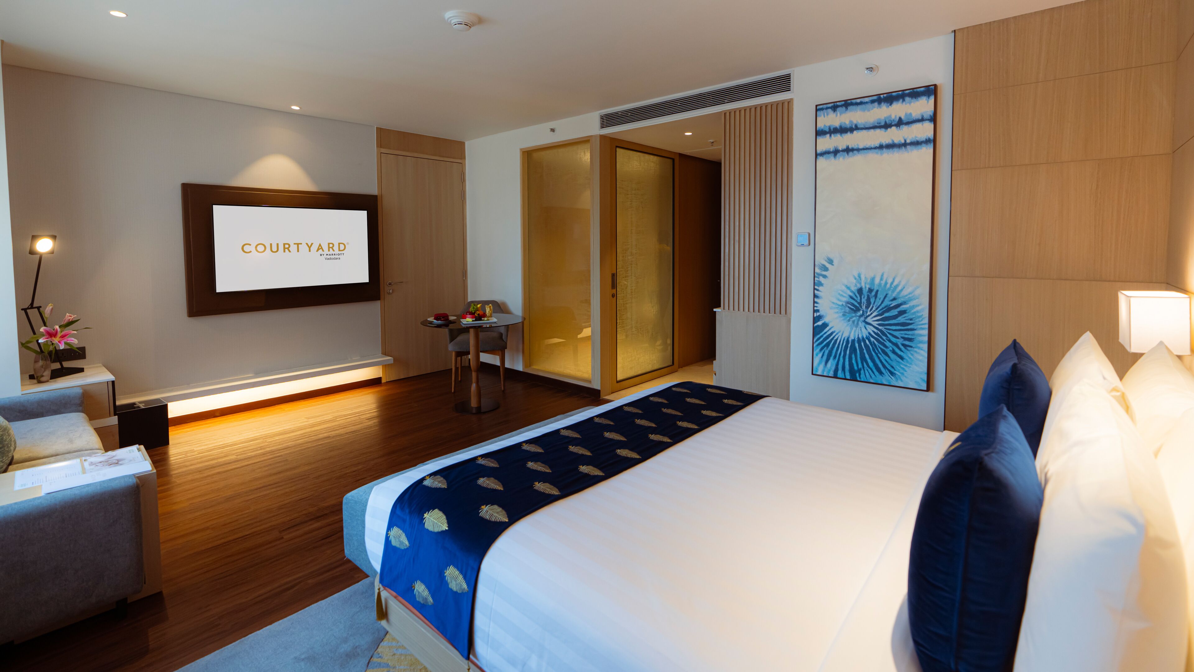 A modern hotel room with a large bed, a desk with a chair, a small table with a vase of flowers, and a sliding door closet.