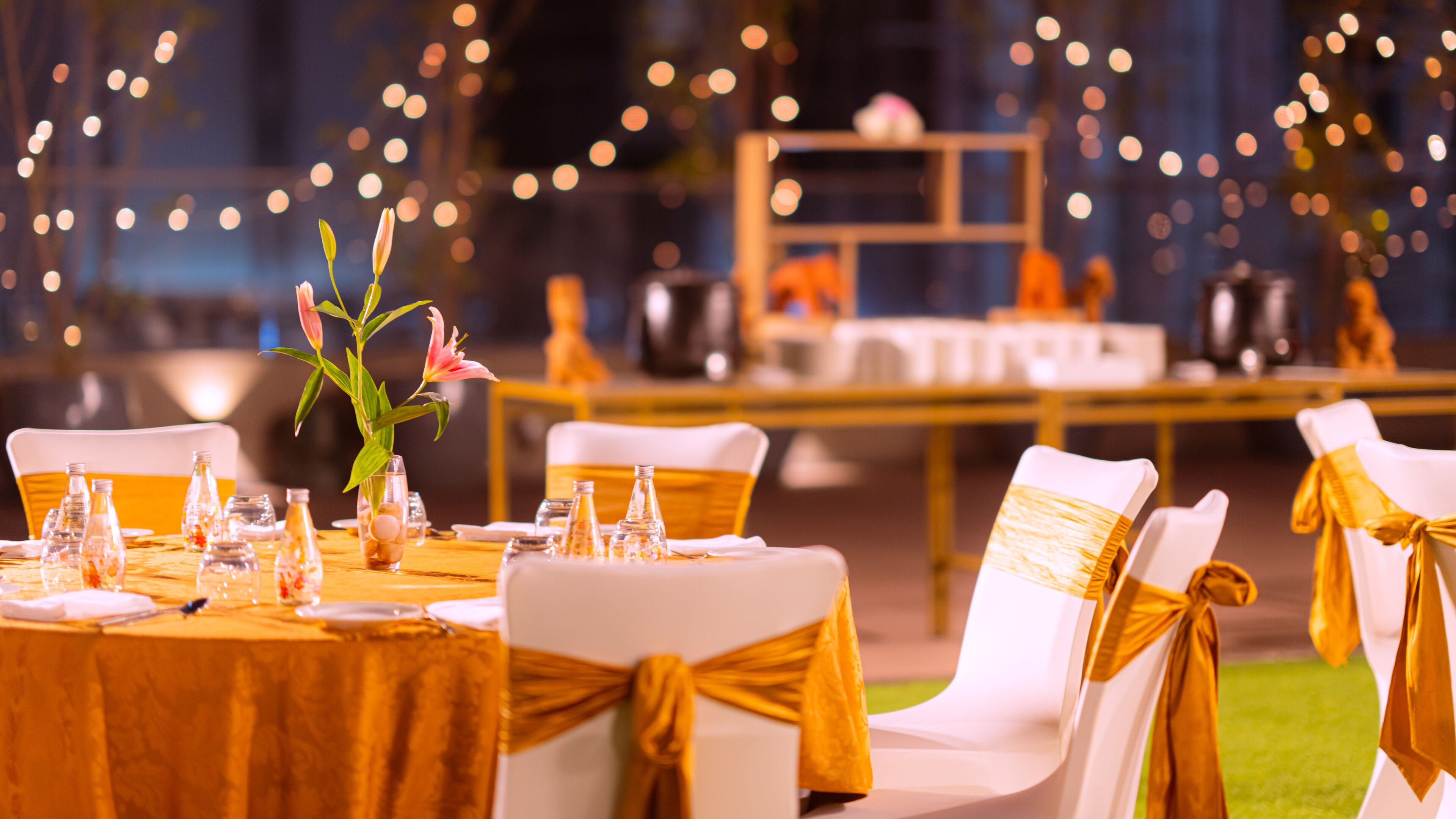 Montaje formal para comedor al aire libre, con sillas y mesas blancas, adornadas con cintas doradas y un centro de mesa floral.