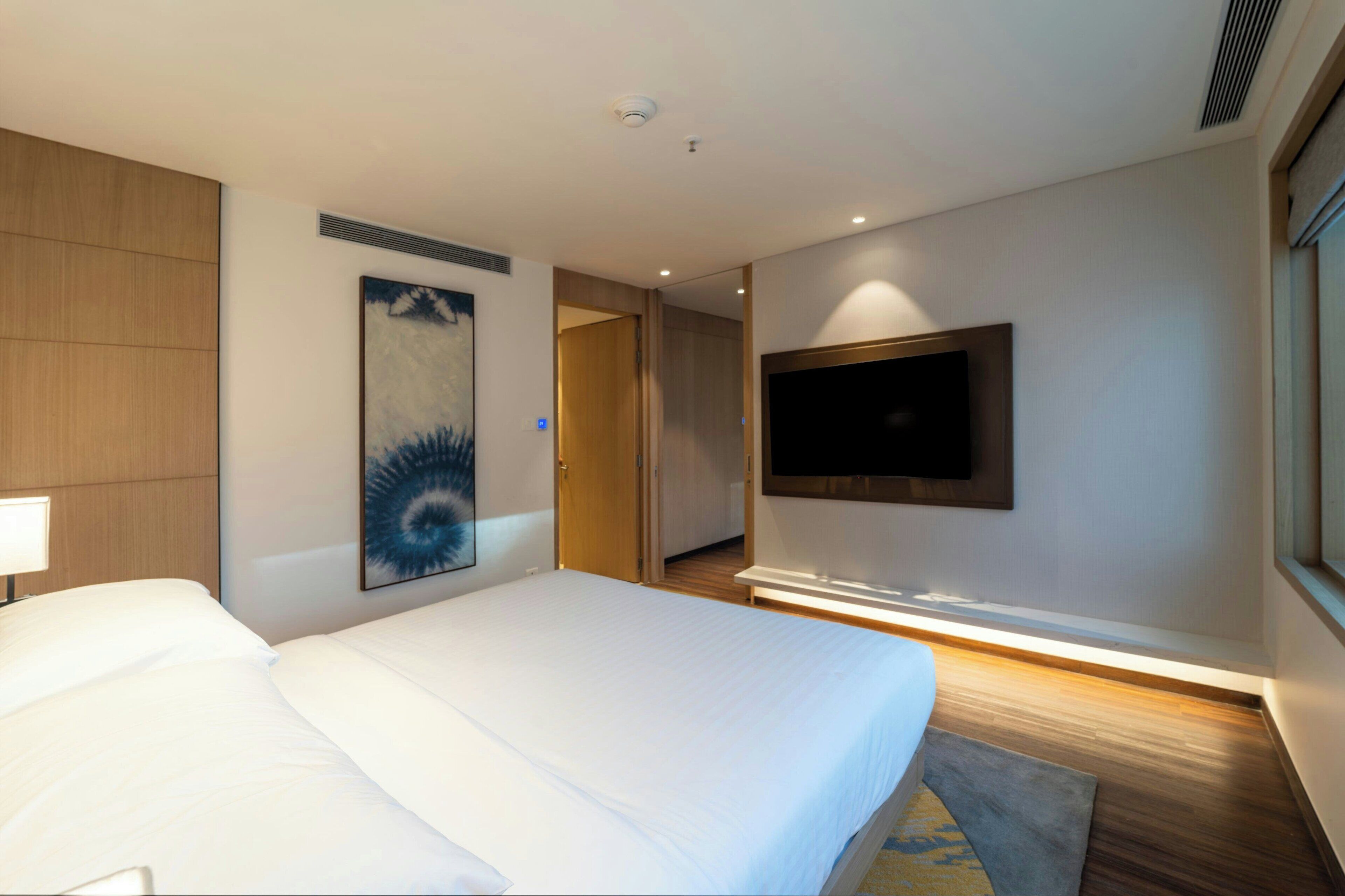 A hotel room with a large bed, a flat-screen TV mounted on the wall, a painting on the wall, and a door leading to another room.