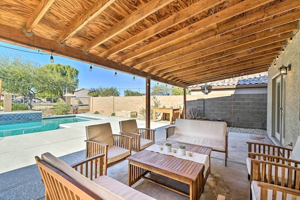 A covered patio area with wooden furniture, a pool, and a stone wall.