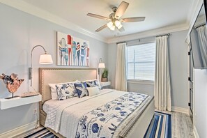 Une chambre à coucher comprenant un lit, un ventilateur de plafond, des œuvres d’art, des lampes de chevet et une fenêtre avec des rideaux.