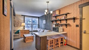 A compact kitchen with a central island, wooden stools, a microwave, and shelves with various items. - Cozy Big Sky Studio ~ 1 Mile to Mtn Village! (Big Sky)