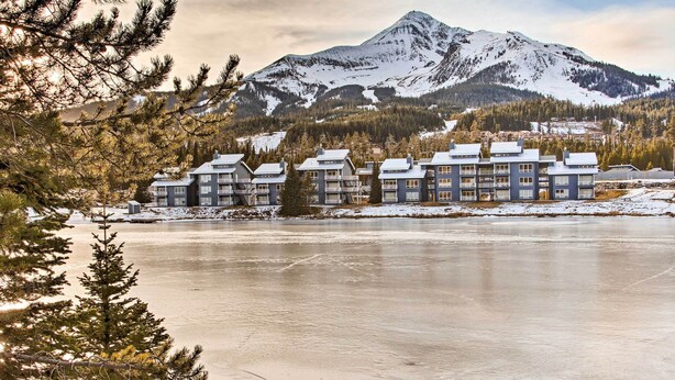 A hotel with multiple floors and balconies, situated by a frozen lake with a mountain in the background. - Cozy Big Sky Studio ~ 1 Mile to Mtn Village! (Big Sky)