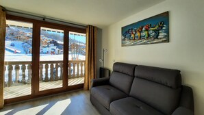 A living room with a dark sofa, a painting, and a view of a snowy outdoor area through large sliding glass doors. - Apartment at the foot of the slopes - 4 guests (Les Belleville)