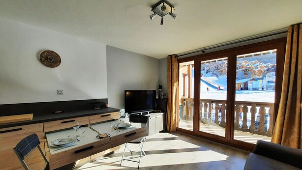 A modern living room with a flat-screen TV, wooden furniture, and a balcony with a view of a ski resort. - Apartment at the foot of the slopes - 4 guests (Les Belleville)