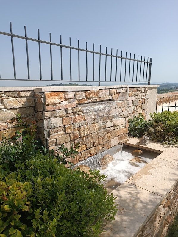 A stone wall with a metal railing, a small waterfall feature, and lush greenery.