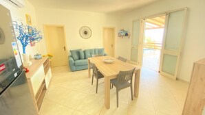 Een ruimtelijke woonruimte met een kitchenette, een eethoek met een houten tafel en stoelen, een blauwe bank en een groot raam met uitzicht.