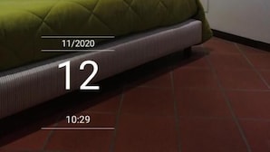 A hotel room with a bed, a wall-mounted lamp, a painting, and a clock displaying the date and time. - Gaia B&b (Castrovillari)
