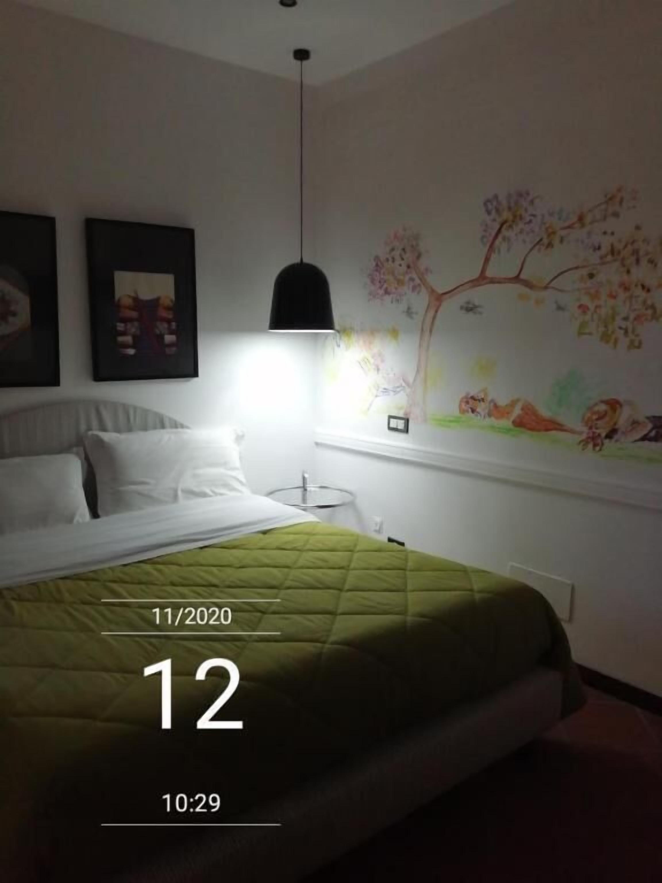 A hotel room with a bed, a wall-mounted painting, a hanging light fixture, and a bedside table.