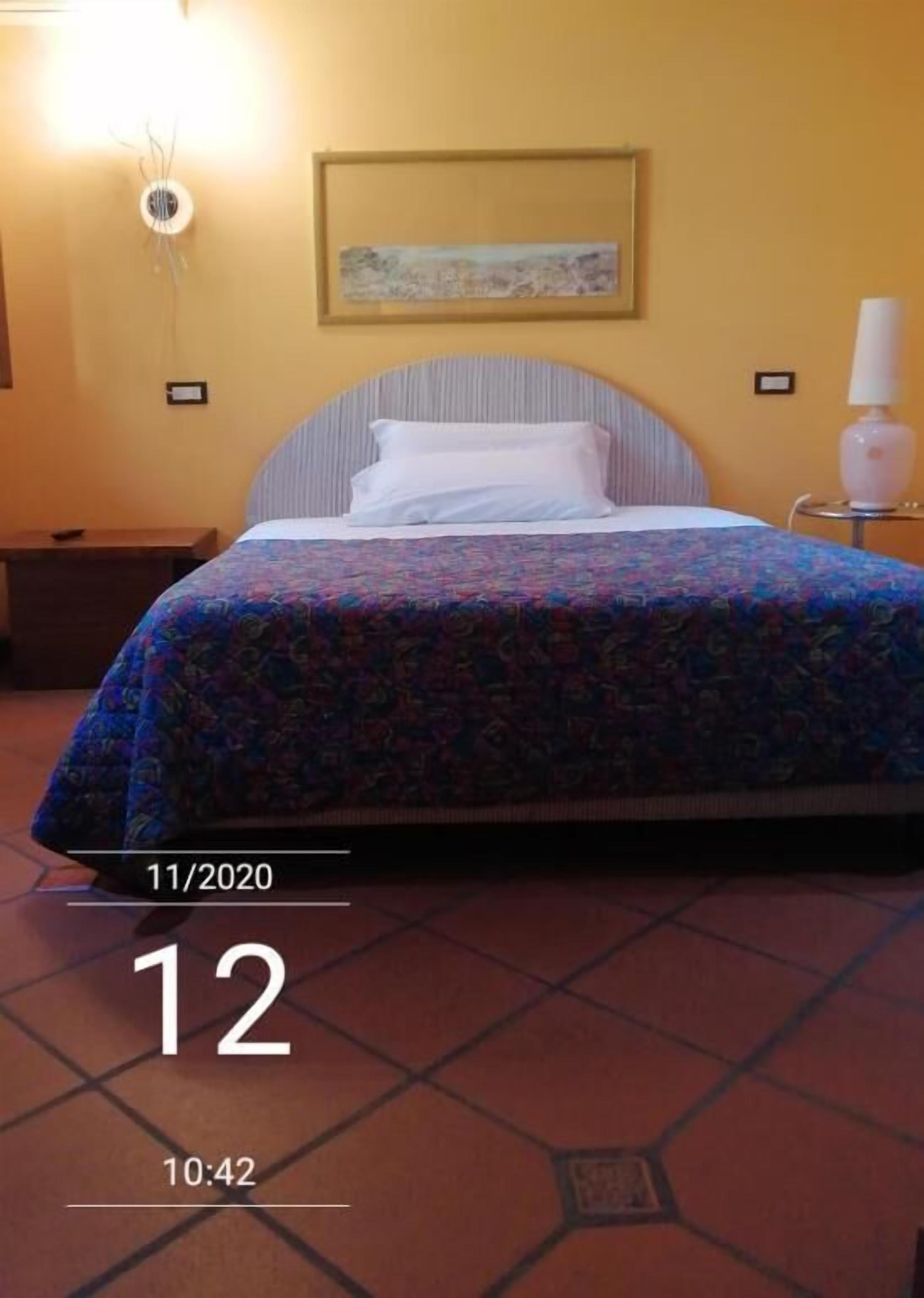 A hotel room with a bed, a nightstand, a wall-mounted lamp, and a painting on the wall.