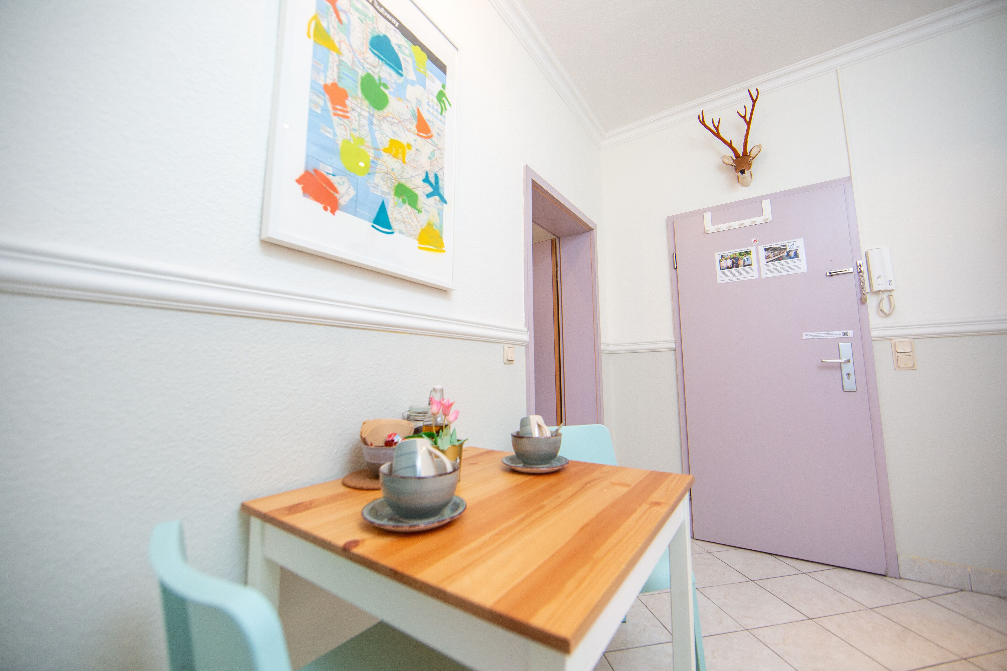 Un couloir avec une table et des chaises en bois, une porte ornée d’une tête de cerf et une carte colorée accrochée au mur.