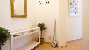 A clean, well-lit hallway with a white wall, a gold-framed mirror, a coat rack, a framed map, a white shelf with magazines, a small potted plant, and a broom leaning against the wall. - ★ Snugly Jugendstil Flat in the ❤ of Erfurt ★ (Erfurt)