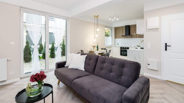 A modern living room with a grey sofa, a black coffee table, and a vase with red flowers. There is a kitchen in the background with white cabinets and a black range hood. - Climatic Studio with Terrace | 500 m to the Old Town Square (Poznan)