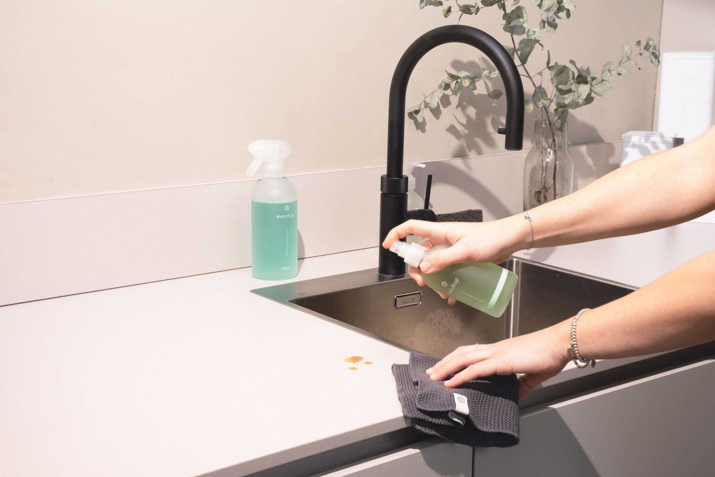 A person cleaning a kitchen sink with a spray bottle and a cloth.