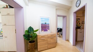 A hallway with a wooden floor, a wall-mounted clock, a framed picture of a field, and a potted plant.