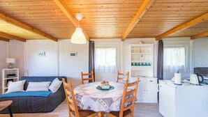 A cozy room with a wooden ceiling, a round table with chairs, a sofa, a small kitchen area, and a window with curtains. - Brittany, Cote de Granit Rose, Chalet with sea view, feet in the water (Trébeurden)