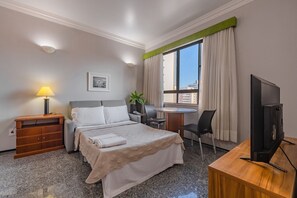 Habitación de hotel con cama, escritorio, silla, televisor, lámpara y ventana con cortinas.