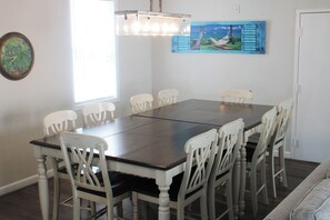 Un comedor con una mesa rectangular rodeada de sillas blancas, un candelabro, un cuadro y un reloj en la pared.