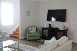 A living room with a green armchair, a beige sofa, a glass coffee table, a dark wooden cabinet, and a flat-screen TV mounted on the wall.