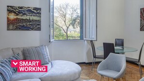 Ein modernes Wohnzimmer mit einer Couch, einem Glastisch und zwei Stühlen. Ein großes Fenster bietet einen Blick ins Freie.