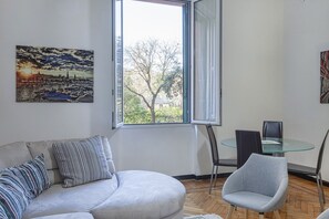 Ein modernes Wohnzimmer mit einer Couch, einem Glastisch und einem Sessel, ein großes Fenster mit Blick auf Bäume und Gebäude sowie ein Gemälde an der Wand.