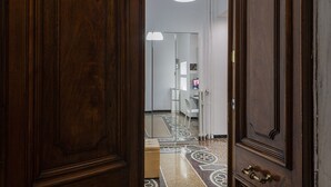 Eine Holztür führt zu einem Flur mit einem gemusterten Boden, einer weißen Deckenleuchte und einem Blick in einen Raum mit Schreibtisch und Stuhl.