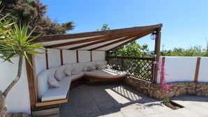 Un espace extérieur aménagé, abrité et doté d’un banc incurvé et de coussins, entouré d’un mur en pierre et d’une pergola en bois.