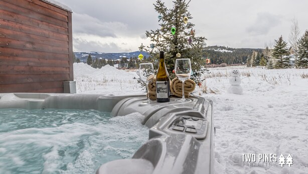 A hot tub with a bottle of wine and glasses, surrounded by snow and a decorated Christmas tree. - Inviting Spanish Peaks Townhome|Private Hot Tub| 15 min Drive to Resort (Big Sky)